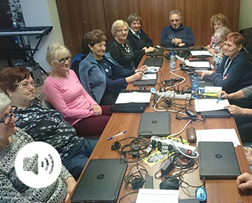 Fotografia przedstawia seniorów siedzacych przy stole, oczekujących na rozpoczęcie szkolenia komputerowego. Zdjęcie pochodzi z archiwum beneficjenta.