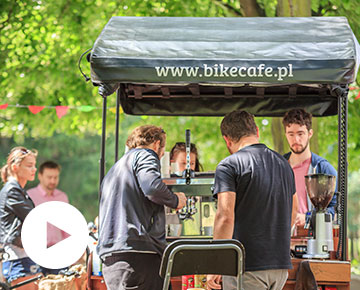 Na zdjęciu: Rower z dystrybutorem do kawy stoi w otoczeniu zieleni. Barista obsługuje dwóch klientów