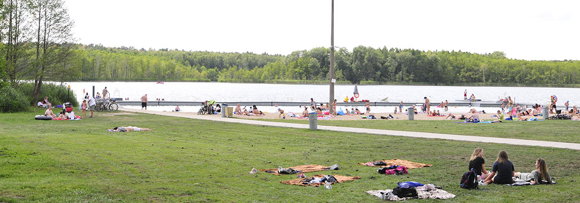 Na zdjęciu ludzie wypoczywający na plaży nad jeziorem w słoneczną pogodę. Autorem zdjęcia jest Jarosław Tomaszewski.