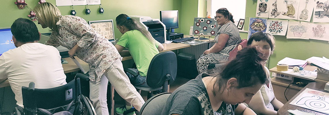 Na zdjęciu uczestnicy projektu w sali pracują przy komputerach. Jedna osoba porusza się na wózku inwalidzkim. Zdjęcie pochodzi z archiwum beneficjenta.