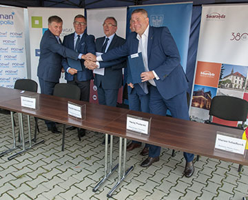 Fotografia przedstawia oficjeli podpisujących umowę, którzy wymieniają uściski dłoni. Autorem zdjęcia jest Jarosław Tomaszewski.