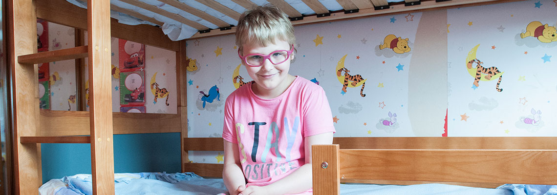 Fotografia przedstawia uśmiechniętą dziewczynkę siedzącą na łóżku. Autorem zdjęcia jest Jarosław Tomaszewski.