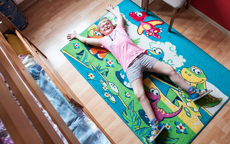 Fotografia przedstawia radosną dziewczynkę leżącą na kolorowym dywanie. Autorem zdjęcia jest Jarosław Tomaszewski.