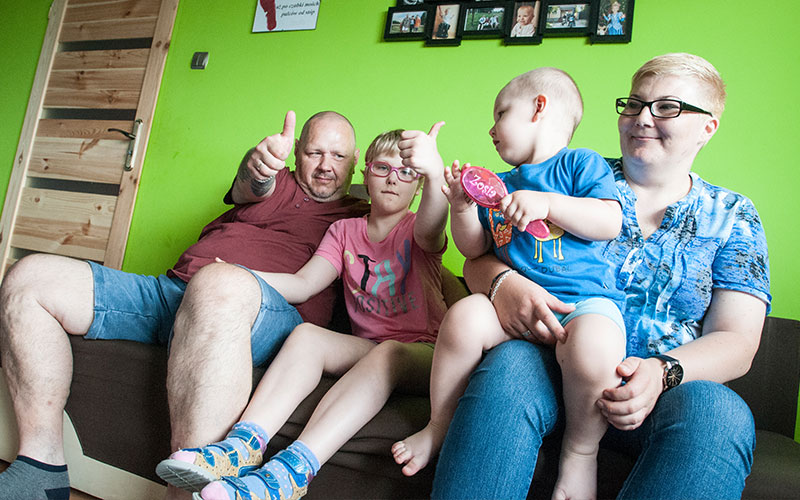 Fotografia przedstawia wesołą, czteroosobową rodzinę siedzącą na kanapie. Autorem zdjęcia jest Jarosław Tomaszewski.