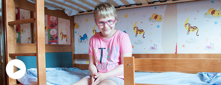 Fotografia przedstawia uśmiechniętą dziewczynkę siedzącą na łóżku. Autorem zdjęcia jest Jarosław Tomaszewski.