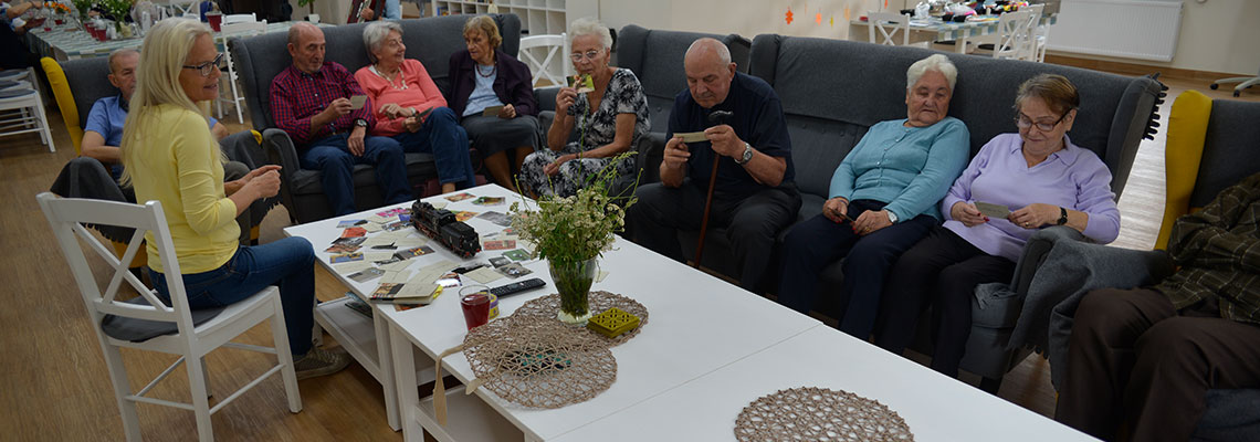 Fotografia przedstawia grupę seniorów siedzących na kanapach, przy stole, rozmawiających z psychologiem, w Domu Seniora. Autorem zdjęcia jest Dominik Wójcik.