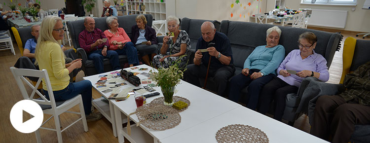 Fotografia przedstawia grupę seniorów siedzących na kanapach, przy stole, rozmawiających z psychologiem, w Domu Seniora. Autorem zdjęcia jest Dominik Wójcik.