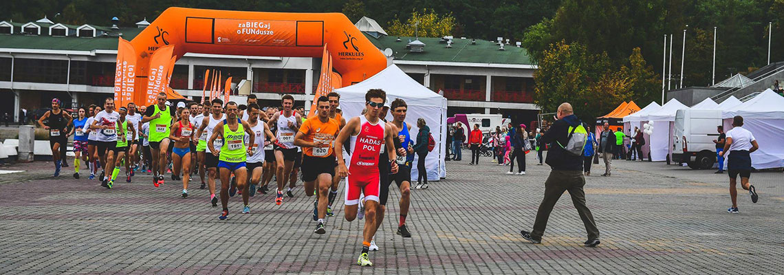 Fotografia przedstawia kilkudziesięciu biegaczy w kolorowych strojach, którzy rozpoczęli wyścig z miejsca startowego. Autorem zdjęcia jest firma Sun and More.