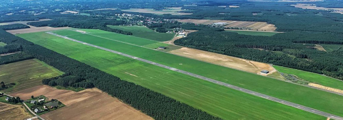 Fotografia wykonana jest z lotu ptaka, przedstawia pas startowy na lotnisku w Kąkolewie, na dalszym planie lasy, pola i zabudowania. Autorem zdjęcia jest Artur Haładyn.