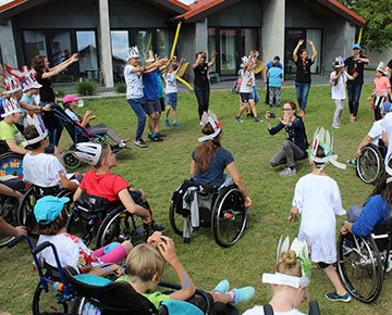 Fotografia przedstawia grupę osób z niepełnosprawnościami, na wózkach inwalidzkich oraz ich opiekunów, we wspólnej zabawie w kole, na trawniku. Zdjęcie pochodzi z archiwum beneficjenta.