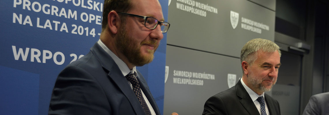 Fotografia przedstawia dwóch mężczyzn w garniturach, prezentujących podpisaną umowę, na tle niebieskiej ścianki z napisem „Wielkopolski Regionalny Program Operacyjny na lata 2014-2020”. Autorem zdjęcia jest Łukasz Karkoszka.