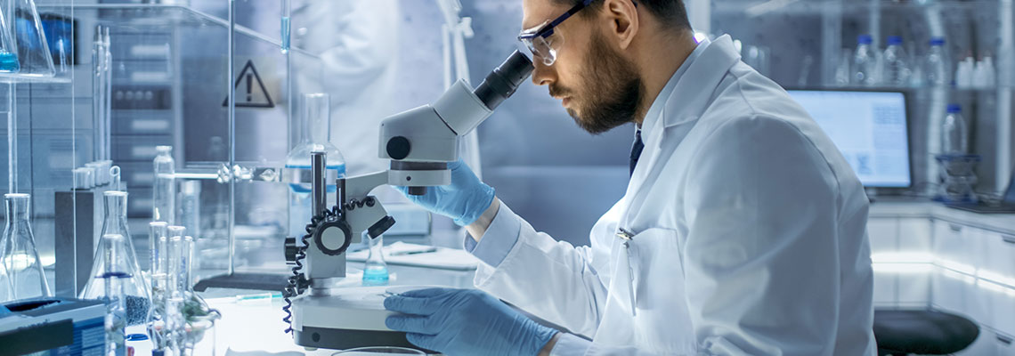 Fotografia przedstawia naukowca w białym fartuchu, w laboratorium. Mężczyzna bada próbkę pod mikroskopem. Zdjęcie pochodzi z  Obrazy licencjonowane przez Gorodenkoff/Shutterstock.com.
