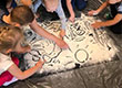 Fotografia przedstawia grupkę dzieci, które rysują palcami wzorki w rozsypanej mące. Zdjęcie pochodzi z archiwum beneficjenta.