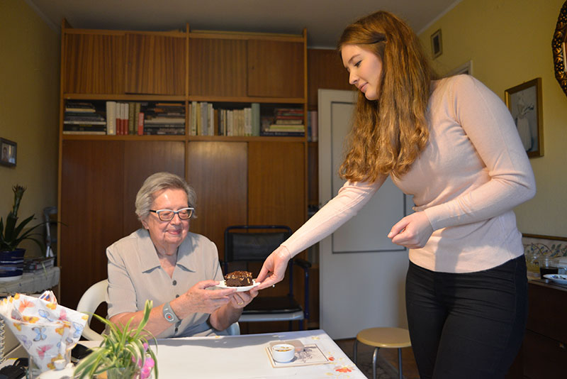 Fotografia przedstawia Nataszę, która stojąc przy stole, podaje siedzącej pani Ewie talerzyk z kawałkiem ciasta. Autorem zdjęcia jest Dominik Wójcik.