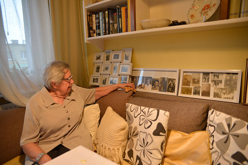 Fotografia przedstawia panią Ewę siedzącą na kanapie. Na oparciu ustawione są ramki z czarno-białymi zdjęciami, przedstawiającymi jej rodzinę. Autorem zdjęcia jest Dominik Wójcik.