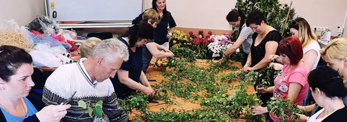 Fotografia przedstawia zajęcia podczas kursu florystycznego. Uczestnicy stoją wzdłuż długiego stołu, na którym rozłożone są zielone rośliny, z których mają stworzyć kompozycje. Zdjęcie pochodzi z archiwum beneficjenta.