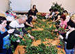 Fotografia przedstawia zajęcia podczas kursu florystycznego. Uczestnicy stoją wzdłuż długiego stołu, na którym rozłożone są zielone rośliny, z których mają stworzyć kompozycje. Zdjęcie pochodzi z archiwum beneficjenta.