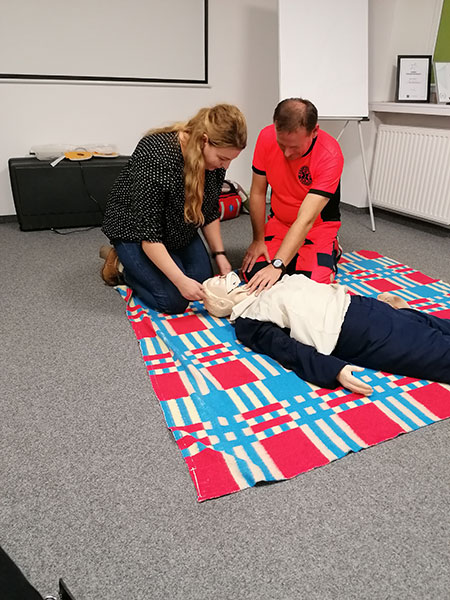Fotografia przedstawia szkolenie z pierwszej pomocy. Kobieta ćwiczy resuscytację na fantomie, instruowana przez ratownika. Oboje klęczą na kolorowym dywanie. Zdjęcie pochodzi z archiwum beneficjenta.