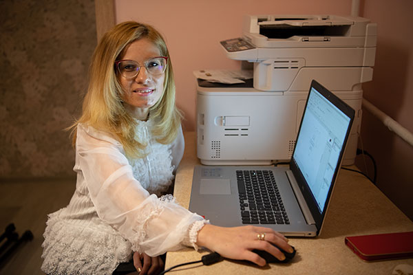 Fotografia przedstawia kobietę o blond włosach, z okularami, która siedzi przy biurku i pracuje na laptopie. Obok, na biurku, stoi urządzenie wielofunkcyjne do drukowania, skanowania i kserowania. Kobieta patrzy w obiektyw aparatu i uśmiecha się. Autorem zdjęcia jest Maciej Motylewski.