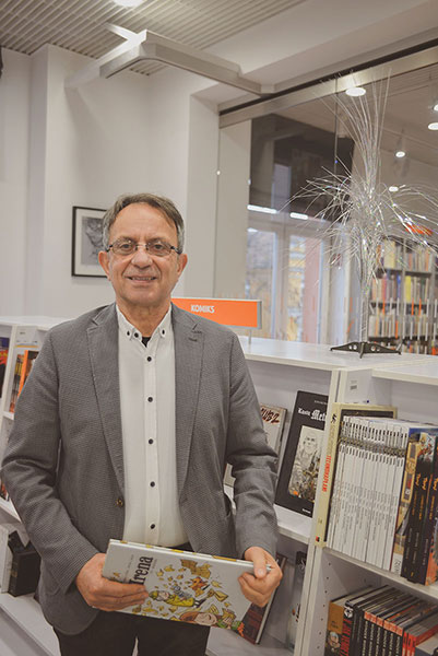 Fotografia przedstawia Włodzimierza Grabowskiego, dyrektora biblioteki. Mężczyzna, w okularach, siwych włosach i szarej marynarce, uśmiecha się, pozując na tle niewysokiego regału z książkami. W ręce trzyma książkę. Autorem zdjęcia jest Łukasz Karkoszka.
