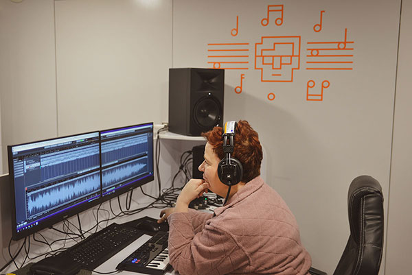 Fotografia przedstawia kobietę obsługującą komputer i specjalistyczne oprogramowanie w studio nagrań. Na głowie ma słuchawki. Autorem zdjęcia jest Łukasz Karkoszka.