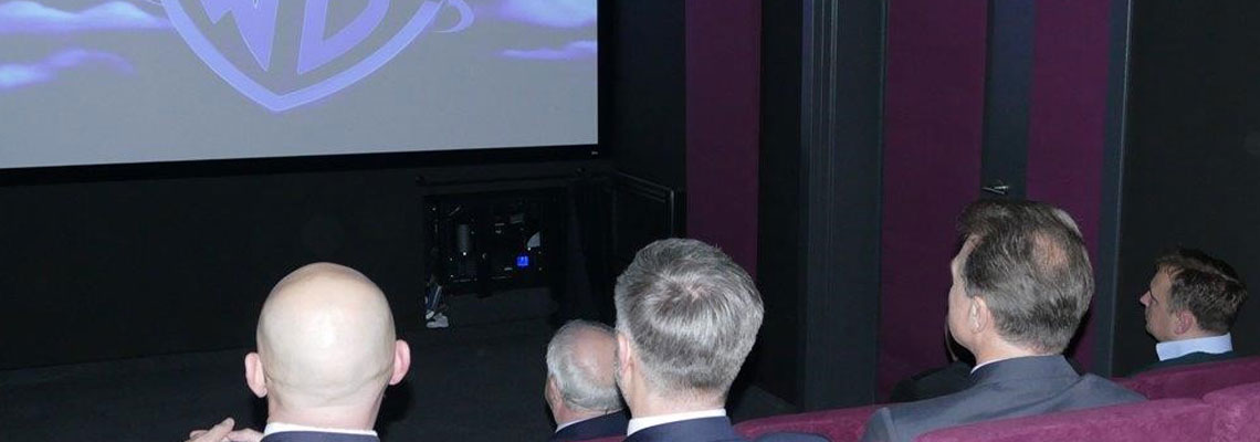 Fotografia przedstawia niewielką salę kinową. Zdjęcie robione jest z tyłu, widać ekran, na którym wyświetlone jest logo Warner Bros, oraz głowy osób siedzących na publiczności. Zdjęcie pochodzi z archiwum beneficjenta.