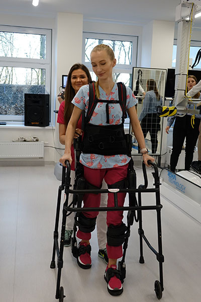Fotografia przedstawia młodą dziewczynę, która ćwiczy za pomocą specjalnego egzoszkieletu i chodzika. Pomaga jej fizjoterapeutka, która asekuruje ją, stojąc z tyłu. Zdjęcie pochodzi z archiwum beneficjenta.
