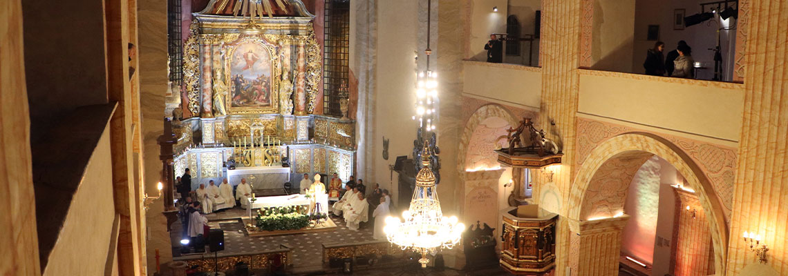 Fotografia przedstawia wnętrze kościoła podczas nabożeństwa. Zdjęcie wykonane jest z balkonu, po lewej stronie nawy głównej. Widoczne są bogate zdobienia prezbiterium. W ławkach siedzą wierni, a przy stoją ołtarzu kapłani. Fotografia pochodzi z archiwum beneficjenta.