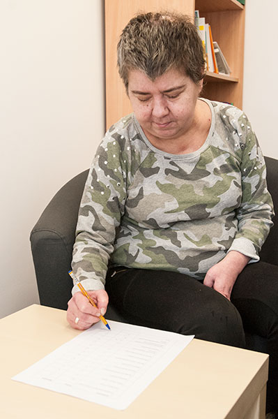 Fotografia przedstawia kobietę w krótkich włosach, siedzącą przy stoliku i wypełniającą dokumenty. Autorem zdjęcia jest Jarosław Tomaszewski.