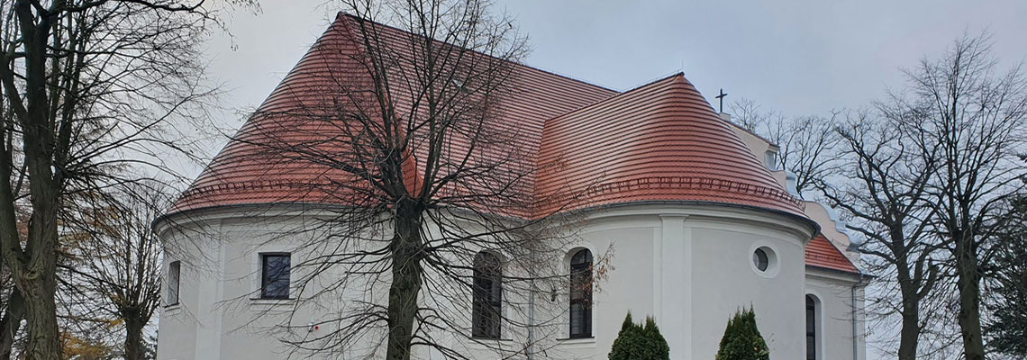 Fotografia przedstawia niewielki kościół o owalnych, białych ścianach i czerwonym, spadzistym dachu. Wokół widoczne są choinki, skalniaki, a po prawej niewielkie oczko wodne. Zdjęcie pochodzi z archiwum beneficjenta.