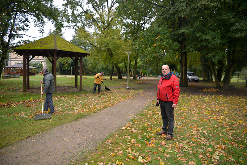 Fotografia przedstawia mężczyznę z ogoloną głową, w czerwonej kurtce, który stoi na trawie przy parkowej alejce. Po drugiej stronie alejki widoczne są dwie osoby, które zgrabiają liście. Autorem zdjęcia jest Dominik Wójcik.