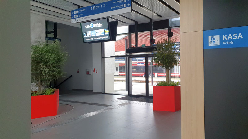 Fotografia przedstawia hol dworca kolejowego w Wolsztynie. Widoczne są przeszklone drzwi, dwie duże czerwone donice z kwiatami, a pod sufitem elektroniczna tablica informacyjna. Zdjęcie pochodzi z archiwum Urzędu Miejskiego w Wolsztynie.
