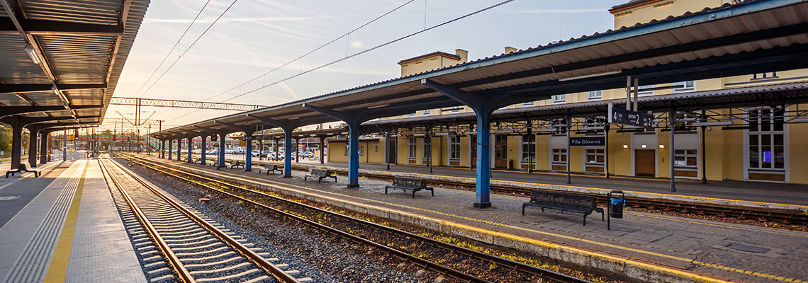 Fotografia przedstawia odnowione, zadaszone perony kolejowe w Pile oraz dwa tory pomiędzy nimi. W tle widać żółty budynek dworca. Zdjęcie pochodzi z archiwum Urzędu Marszałkowskiego Województwa Wielkopolskiego.