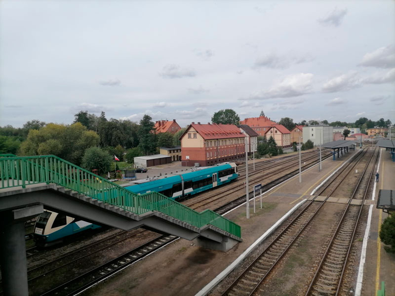 Fotografia wykonana z kładki nad torami, przedstawia stację kolejową. Widać peron i kilka torów z lewej oraz jeden z prawej strony. Widoczny jest wjeżdżający, biało-zielony pociąg. Autorem zdjęcia jest Radosław Śledziński.