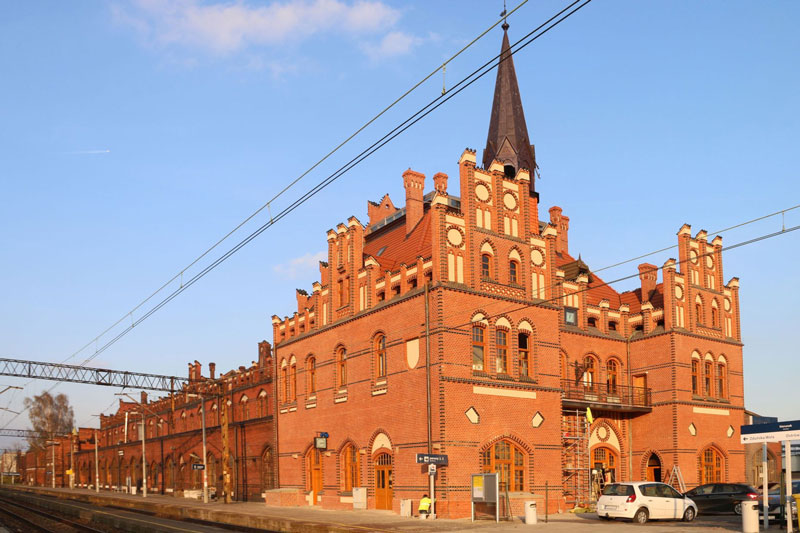 Fotografia przedstawia dworzec kolejowy w Nowych Skalmierzycach. Budynek przypomina wyglądem katedrę, jest z czerwonej cegły, ma strzelistą wieżyczkę. Zdjęcie pochodzi z archiwum Urzędu Miejskiego w Nowych Skalmierzycach.