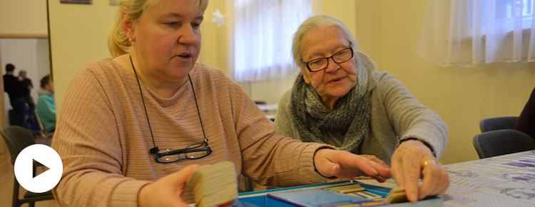 Fotografia przedstawia opiekunkę i seniorkę, które siedzą przy stole i rozkładają karty do gry planszowej. Autorem zdjęcia jest Dominik Wójcik.