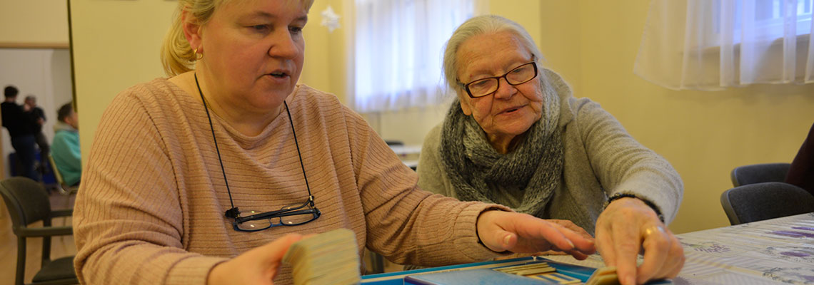 Fotografia przedstawia opiekunkę i seniorkę, które siedzą przy stole i rozkładają karty do gry planszowej. Autorem zdjęcia jest Dominik Wójcik.
