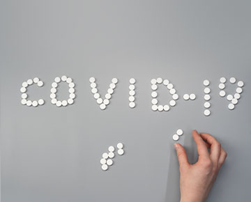 Grafika przedstawia napis „COVID-19” ułożony z okrągłych, białych tabletek na szarym podłożu. U dołu zdjęcia widocznych jest jeszcze kilka innych tabletek oraz dłoń układająca napis. Autorem zdjęcia jest cottonobro.