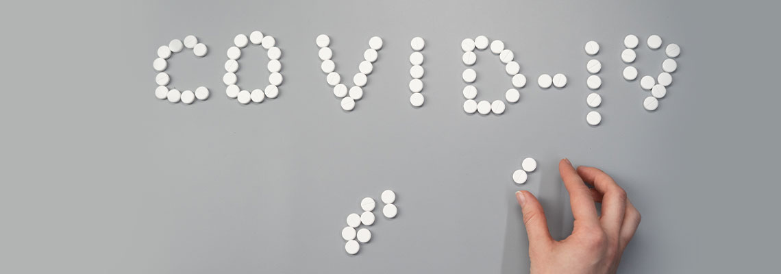 Grafika przedstawia napis „COVID-19” ułożony z okrągłych, białych tabletek na szarym podłożu. U dołu zdjęcia widocznych jest jeszcze kilka innych tabletek oraz dłoń układająca napis. Autorem zdjęcia jest cottonobro.