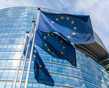Fotografia przedstawia trzy powiewające flagi Unii Europejskiej na tle przeszklonego budynku, prawdopodobnie Parlamentu Europejskiego. Zdjęcie pochodzi z  Obrazy licencjonowane przez Depositphotos.com/Drukarnia Chroma.
