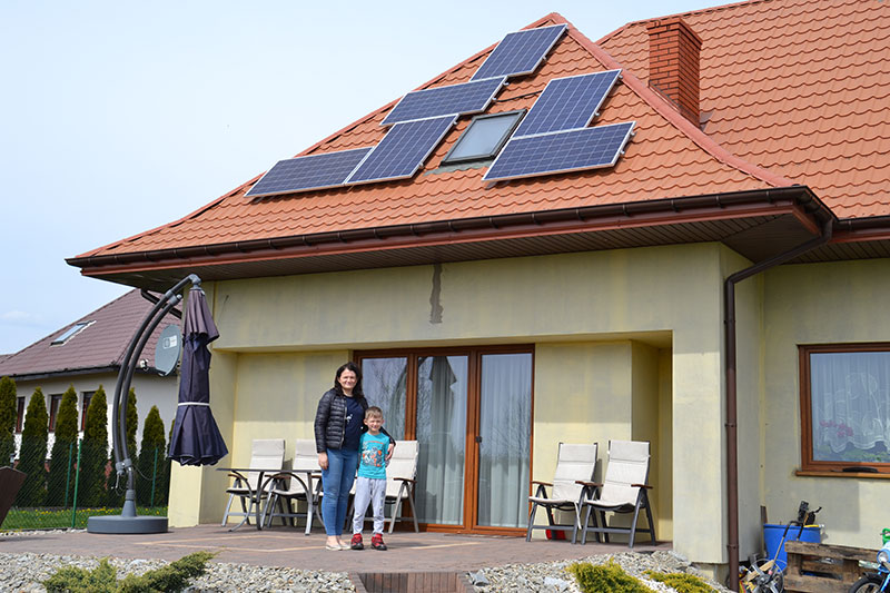 Fotografia przedstawia Joannę Błaszczyk wraz z synek stojącą przed przeszklonymi drzwiami ich domu. Na skośnym dachy krytym dachówką widać kilka paneli słonecznych. Zdjęcie pochodzi z archiwum Gminy Malanów.