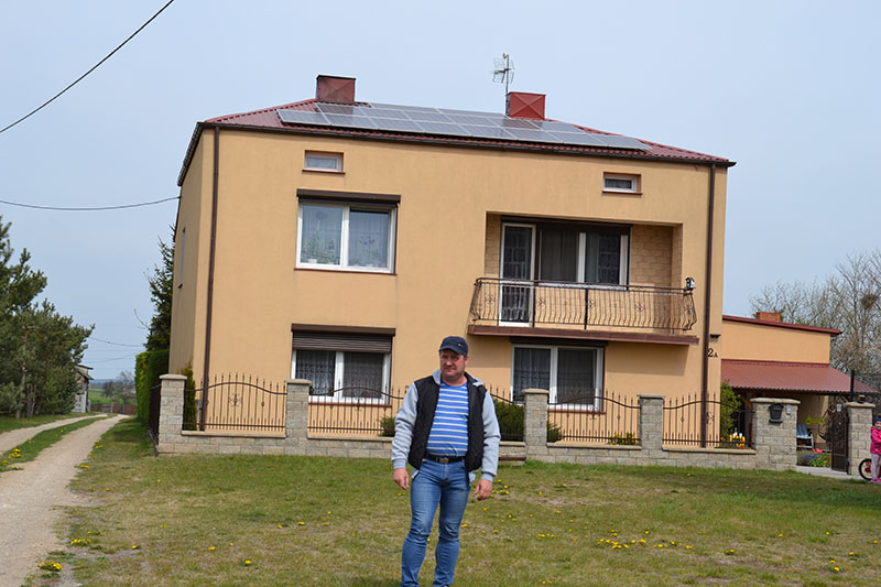 Fotografia przedstawia Macieja Kornackiego – mężczyznę w średnim wieku ubranego w czapkę z daszkiem, kurtkę i dżinsy, który pozuje na tle swojego domu. Na skośnym dachu widoczne są panele słoneczne. Zdjęcie pochodzi z archiwum Gminy Malanów.