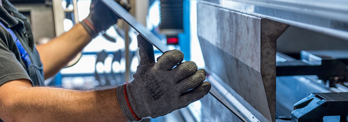 Fotografia przedstawia mężczyznę pracującego przy maszynie. Widoczne są ręce pracownika, jego dłonie są w rękawiczkach. Mężczyzna trzyma przedmiot w kształcie kwadratowej płyty, który jest obrabiany w maszynie. Fotografia została wykonana przez jarmoluk – 143740/canva.pl.