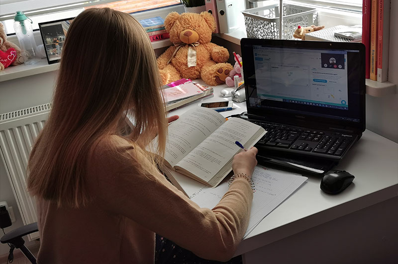 Fotografia przedstawia młodą uczennicę czytającą zadanie w książce, następnie przepisującą dane na kartkę. Na biurku stoi laptop. Na ekranie widoczne jest okienko z połączeniem internetowym z nauczycielem. Zdjęcie pochodzi z archiwum prywatnego.
