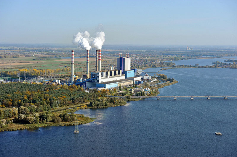 Fotografia przedstawia elektrownię w Koninie z lotu ptaka. Widoczne są bloki elektrowni, cztery wysokie kominy, z dwóch wydobywa się biały dym, oraz ogromny zbiornik wodny. Zdjęcie pochodzi z archiwum Urzędu Miejskiego w Koninie.
