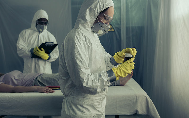 Fotografia przedstawia dwóch pracowników medycznych, prawdopodobnie lekarzy w kombinezonach, maskach i rękawiczkach, przy łóżku pacjenta w izolatce. Jeden z lekarzy zapisuje coś w karcie drugi zabezpiecza próbkę. Zdjęcie pochodzi z Adobe Stock.