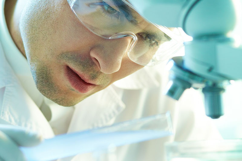 Fotografia przedstawia naukowca w ochronnych okularach i białym fartuchu, który pochyla się nad preparatem pod mikroskopem. Zdjęcie pochodzi z Obrazy licencjonowane przez Depositphotos.com/Drukarnia Chroma.