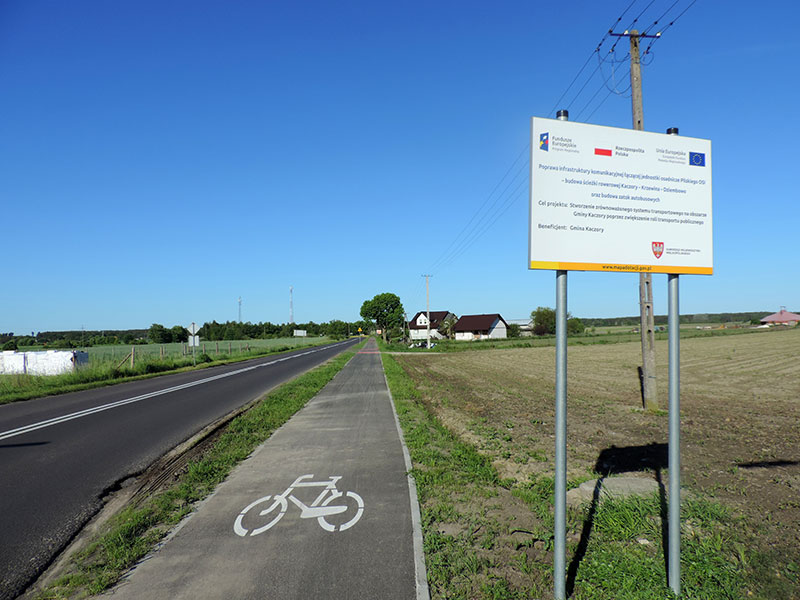 Fotografia przedstawia ścieżkę rowerową, biegnącą wzdłuż drogi przez wiejską okolicę. Na pierwszym planie widoczna jest tablica z informacjami dotyczącymi projektu, w ramach którego powstała ścieżka. Zdjęcie pochodzi z archiwum beneficjenta.