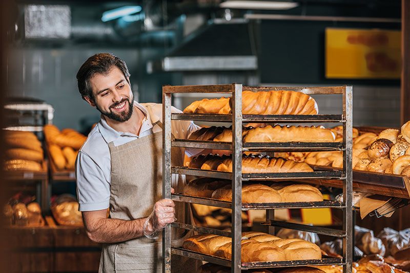 Fotografia przedstawia uśmiechniętego mężczyznę - pracownika piekarni, który przesuwa stojak, na którym leży świeżo upieczony chleb. Wokół widać inne wyroby, poza chlebem także bułki. Zdjęcie pochodzi z Obrazy licencjonowane przez Depositphotos.com/Drukarnia Chroma.