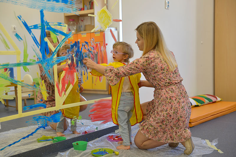 Fotografia przedstawia dziecko w żłobku, w żółtym fartuchu i okularach, które, z pomocą klęczącej za nim opiekunki, maluje dłońmi, kolorowymi farbami, po lustrze. Autorem zdjęcia jest Dominik Wójcik.